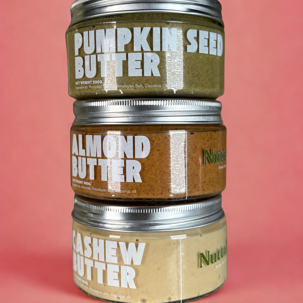 Three jars of different nut butters stacked on a pink background. Healthy Nut Butter by NuttaNut