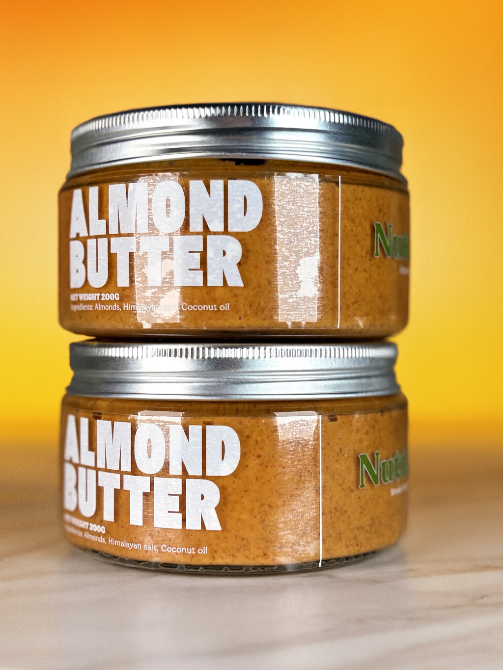 Two jars of almond butter stacked on a wooden surface with a yellow gradient background. NuttaNut Indonesia, best healthy nut and seed butter in Indonesia